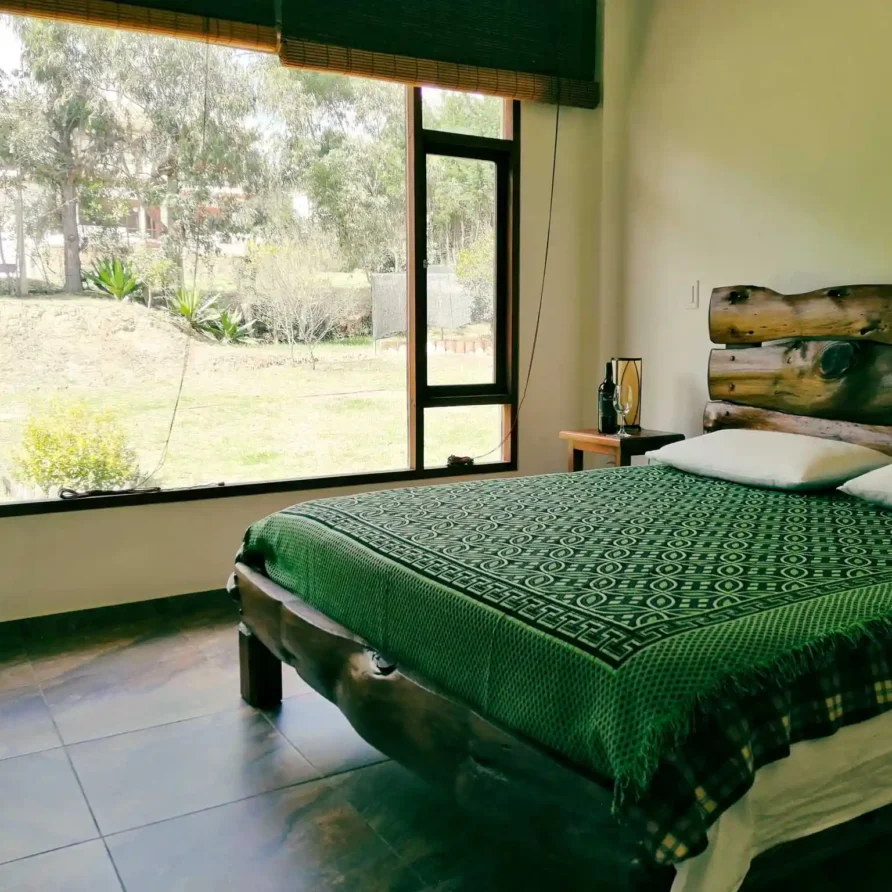 Dormitorio rústico en Casa Zuetana, Zipaquirá, con cama de troncos, manta verde y ventanal al jardín.