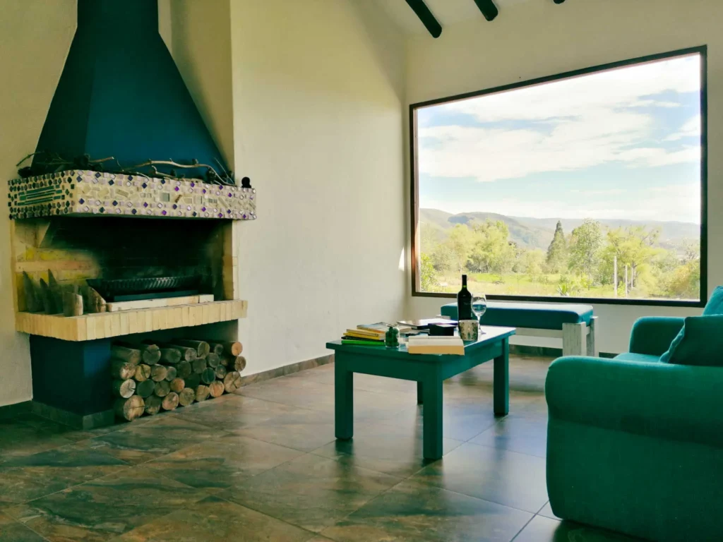 Sala confortable de Casa Zuetana en Zipaquirá con chimenea rústica, sillón turquesa y gran ventanal con vista natural.