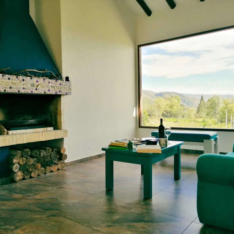 Sala confortable de Casa Zuetana en Zipaquirá con chimenea rústica, sillón turquesa y gran ventanal con vista natural.