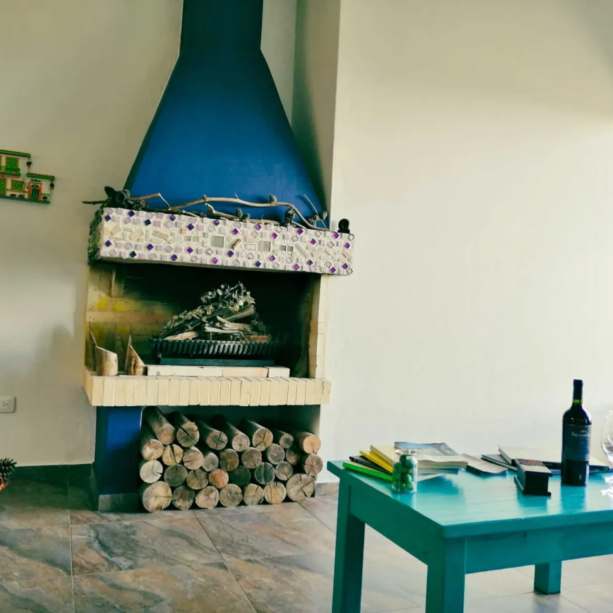 Acogedora sala de Casa Zuetana, Zipaquirá, con chimenea de mosaico azul, mesa de madera y botella de vino.