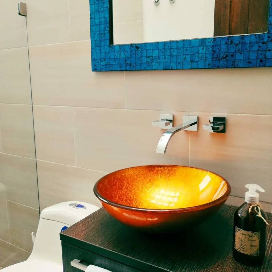 Baño moderno con lavabo de cristal naranja, espejo mosaico azul y grifería de pared en Casa Zuetana, Zipaquirá.