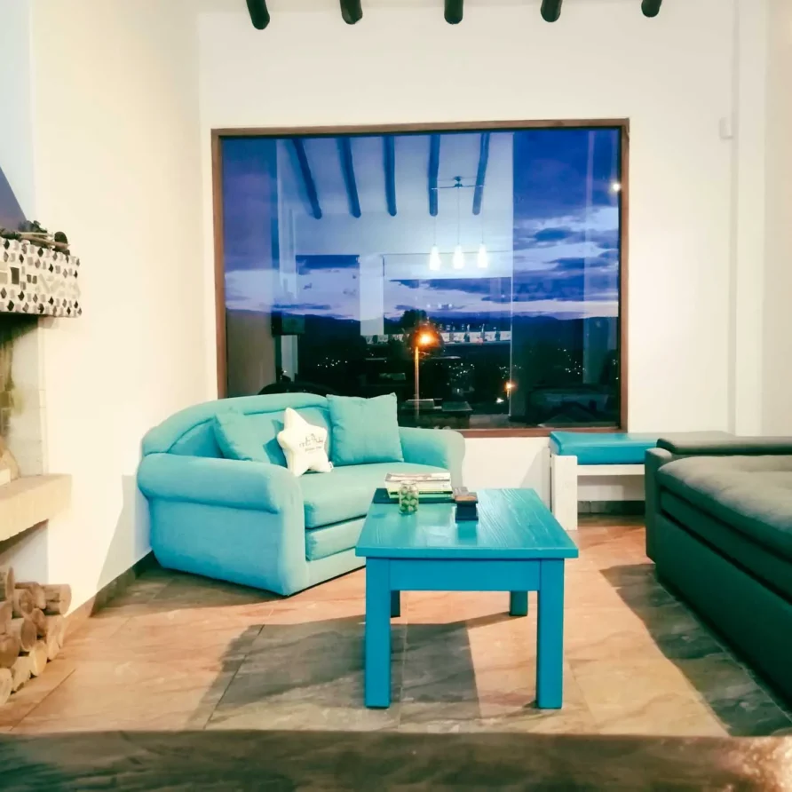 Acogedora sala de estar en Casa Zuetana, Zipaquirá, con sofás turquesa, chimenea rústica y vista nocturna de la ciudad.