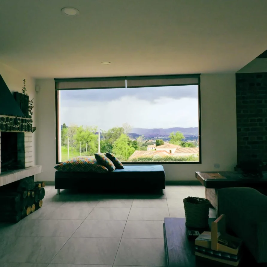 Sala de estar en Casa Hichatana, Zipaquirá, con chimenea, sofá y gran ventanal ofreciendo vistas panorámicas al paisaje.