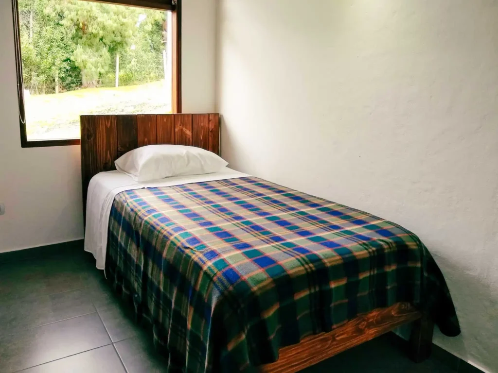 Habitación individual con cama de madera, manta a cuadros y vista a la naturaleza en Casa Hichatana Zipaquirá.
