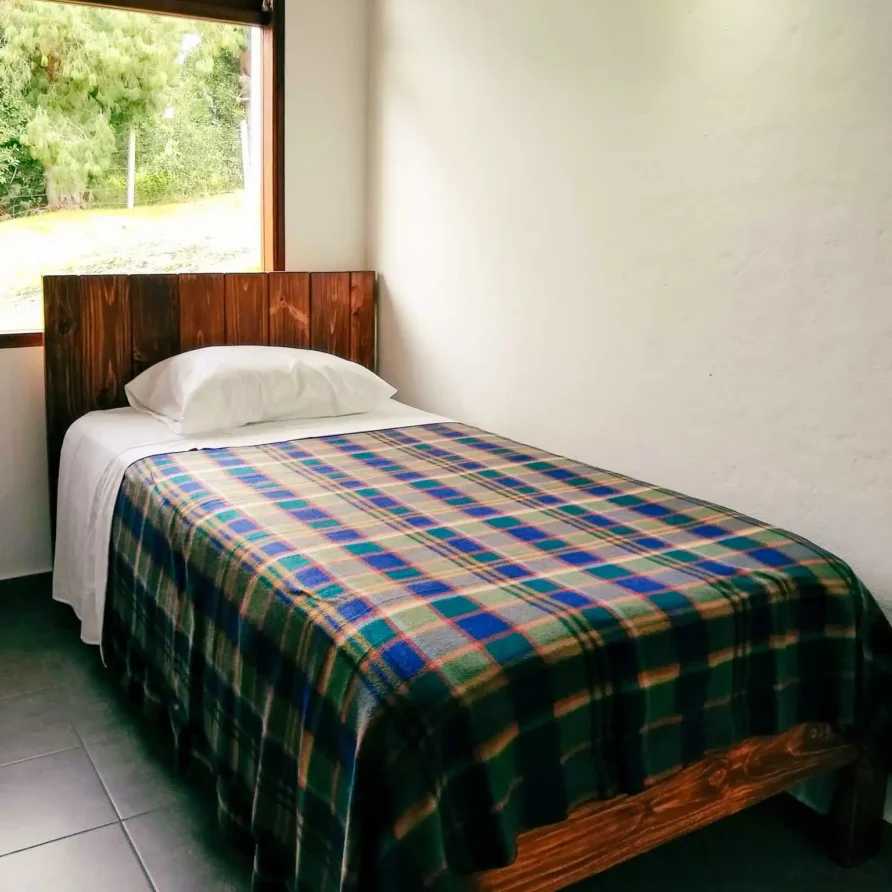 Habitación individual con cama de madera, manta a cuadros y vista a la naturaleza en Casa Hichatana Zipaquirá.