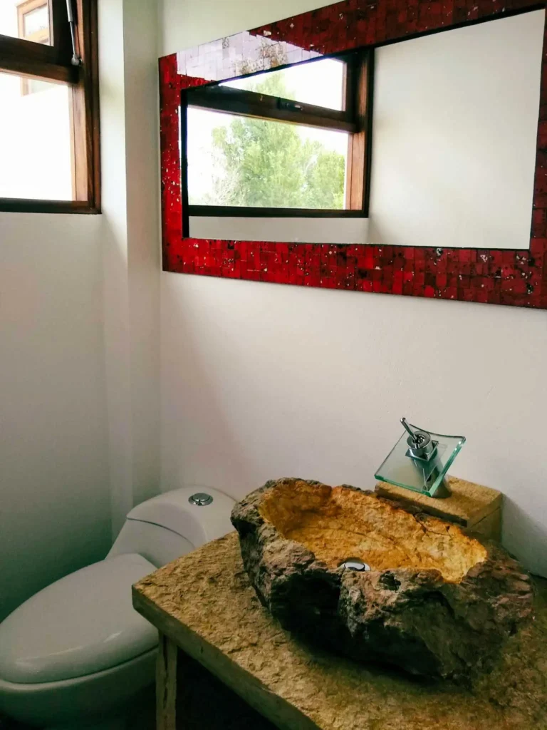 Baño rústico-moderno en Casa Hichatana, Zipaquirá, con lavamanos de piedra y espejo de mosaico rojo.