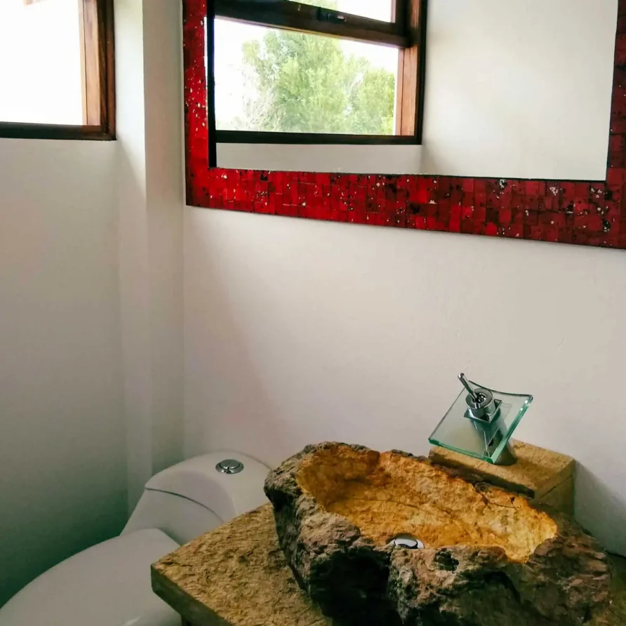 Baño rústico-moderno en Casa Hichatana, Zipaquirá, con lavamanos de piedra y espejo de mosaico rojo.