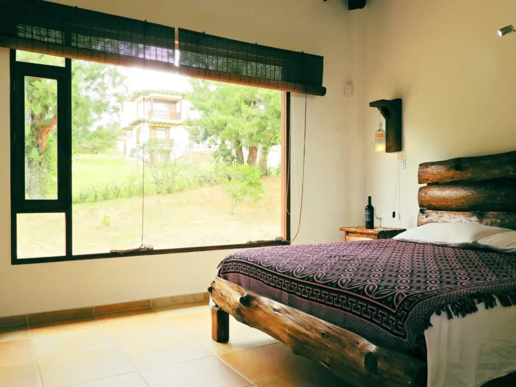 Dormitorio rústico en Casa Hichatana Zipaquirá con cama de troncos de madera y gran ventana con vista al jardín.