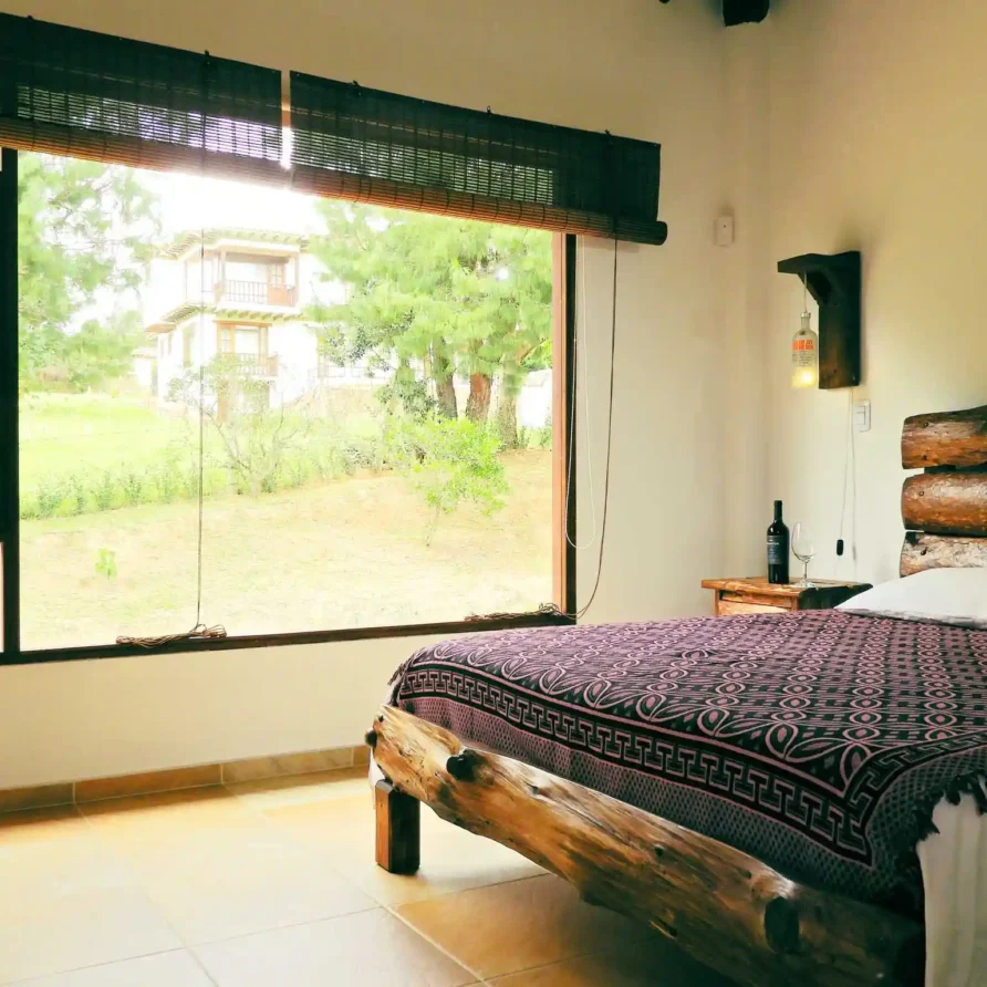 Dormitorio rústico en Casa Hichatana Zipaquirá con cama de troncos de madera y gran ventana con vista al jardín.