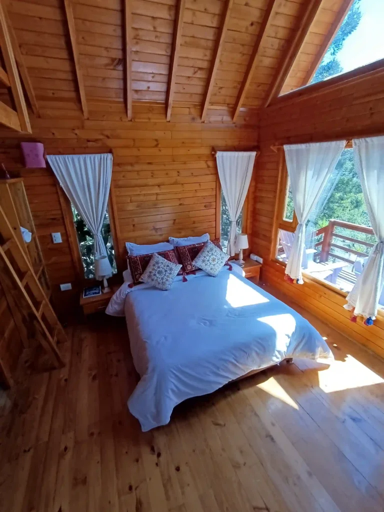 Dormitorio principal de cabaña de madera premium 'El Enueño' con cama, ventanales, luz natural y balcón.