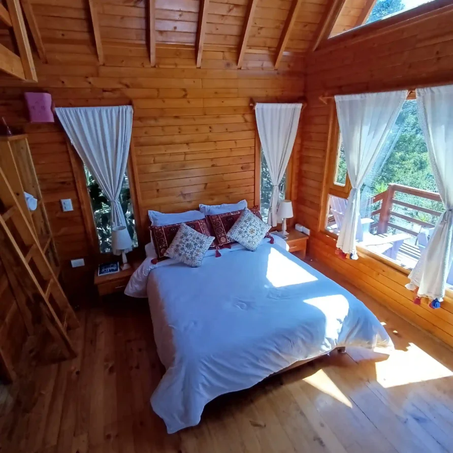 Dormitorio principal de cabaña de madera premium 'El Enueño' con cama, ventanales, luz natural y balcón.