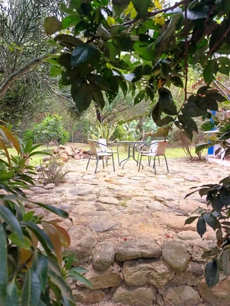 Patio de piedra con mesa y sillas de jardín, rodeado de exuberante vegetación en una cabaña de Villa de Leyva.
