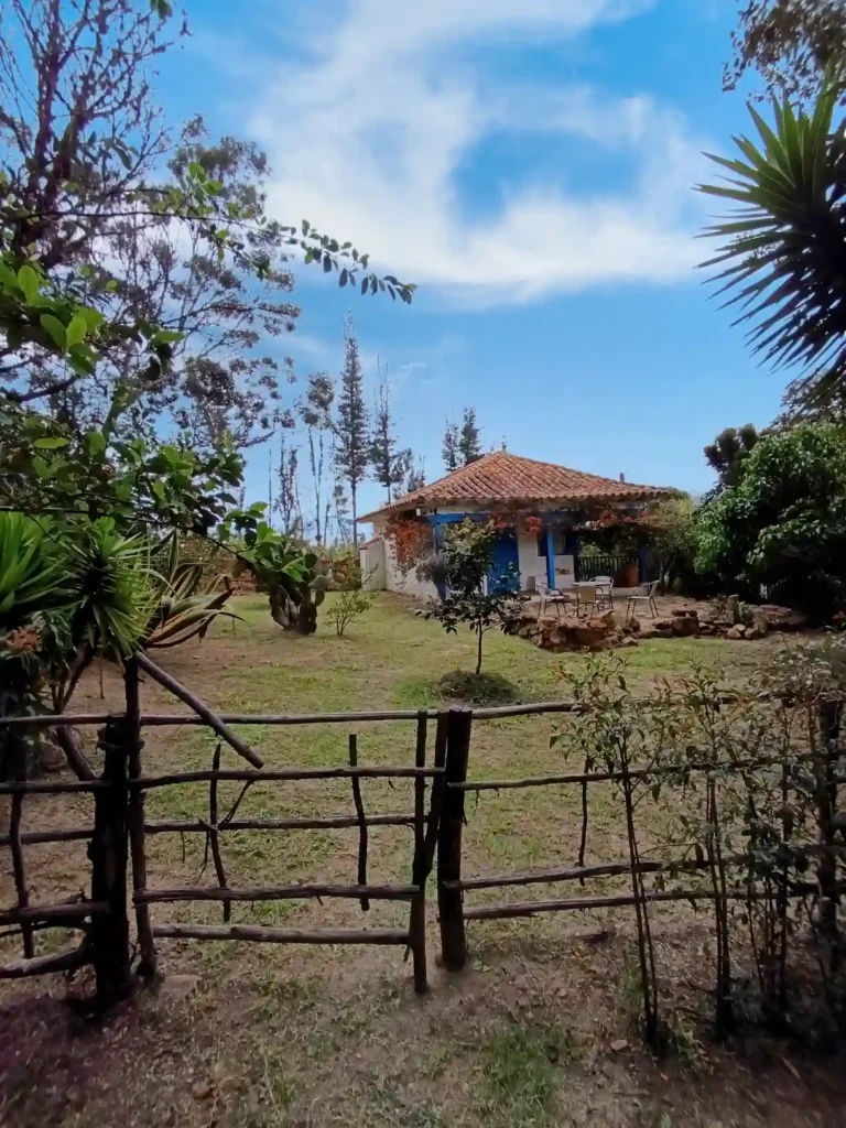 Cabaña rústica El Oteo en Villa de Leyva con techo de teja, puertas azules y un frondoso jardín verde.