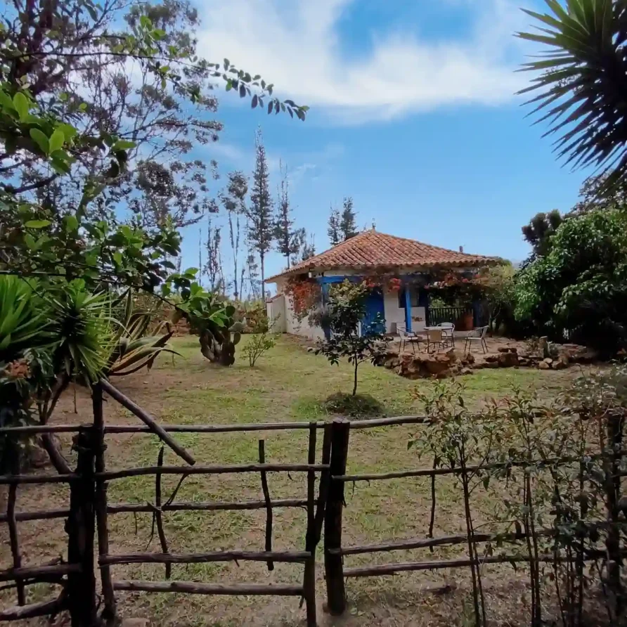 Cabaña rústica El Oteo en Villa de Leyva con techo de teja, puertas azules y un frondoso jardín verde.
