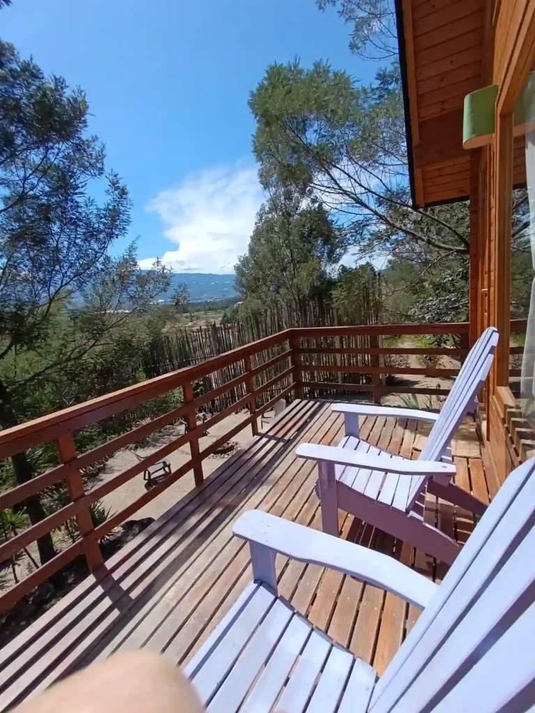 Balcón rústico en hospedaje premium Cabaña el ensueño con barandilla y sillas para relajarse, rodeado de árboles y vista panorámica.
