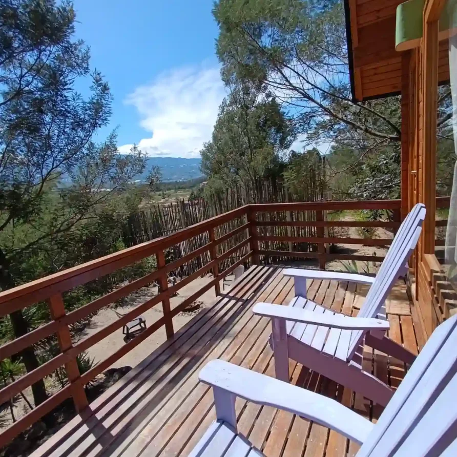Balcón rústico en hospedaje premium Cabaña el ensueño con barandilla y sillas para relajarse, rodeado de árboles y vista panorámica.