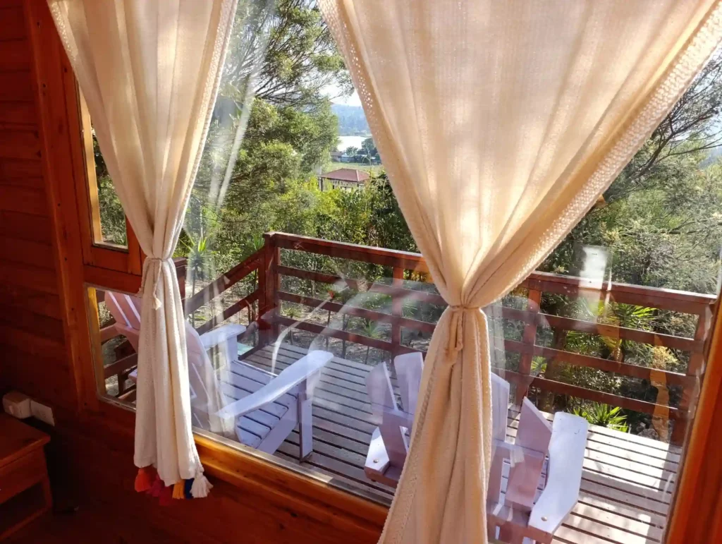 Ventana de cabaña el ensueño en Vereda Monquirá con cortinas claras y panorámica a balcón de madera y entorno natural.
