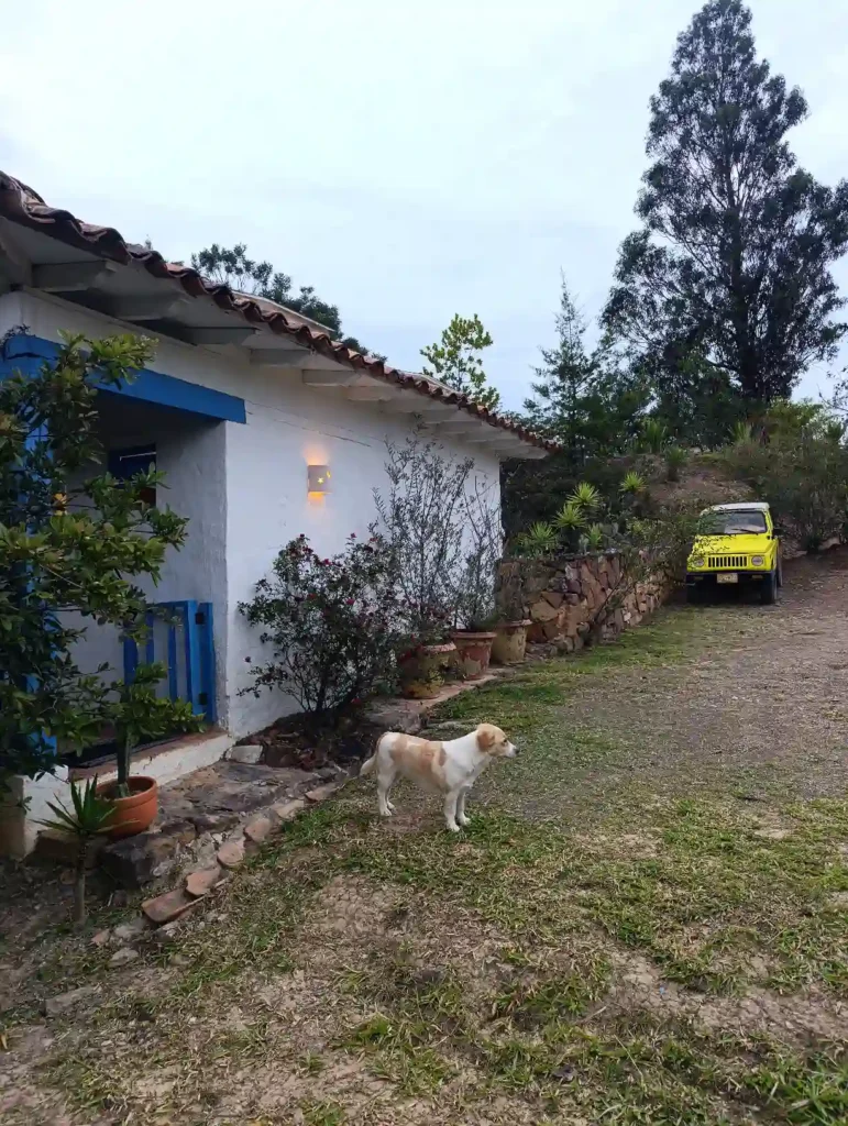 Cabaña blanca El Oteo en Villa de Leyva con perro, jeep amarillo y jardines exuberantes.