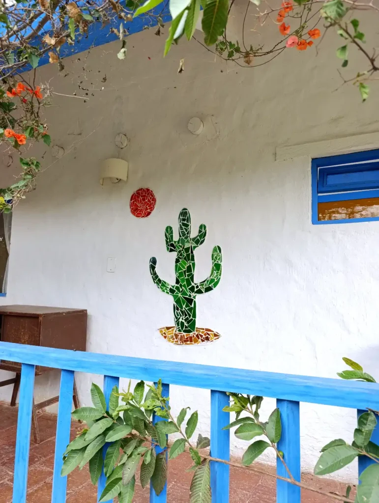 Mosaico de cactus y sol en pared blanca de cabaña en Villa de Leyva, con barandilla azul y vegetación.
