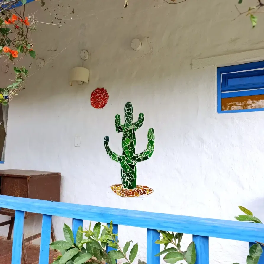 Mosaico de cactus y sol en pared blanca de cabaña en Villa de Leyva, con barandilla azul y vegetación.