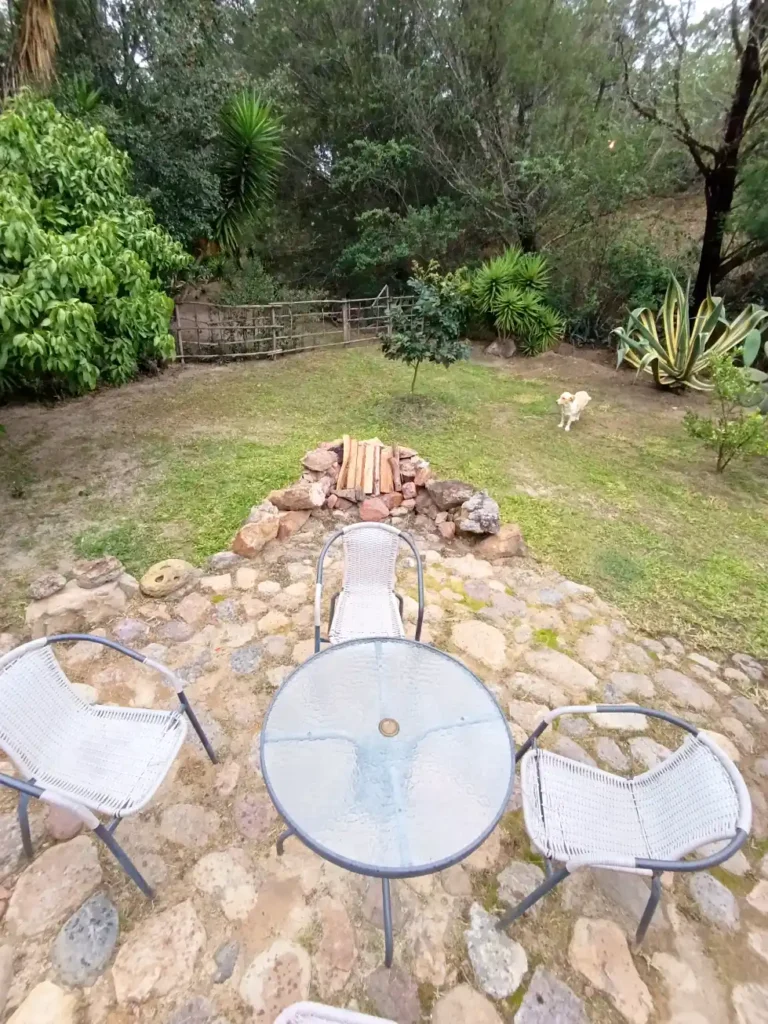Jardín de Cabaña El Oteo en Villa de Leyva con patio de piedra, mesa, sillas, fogata y vegetación natural.