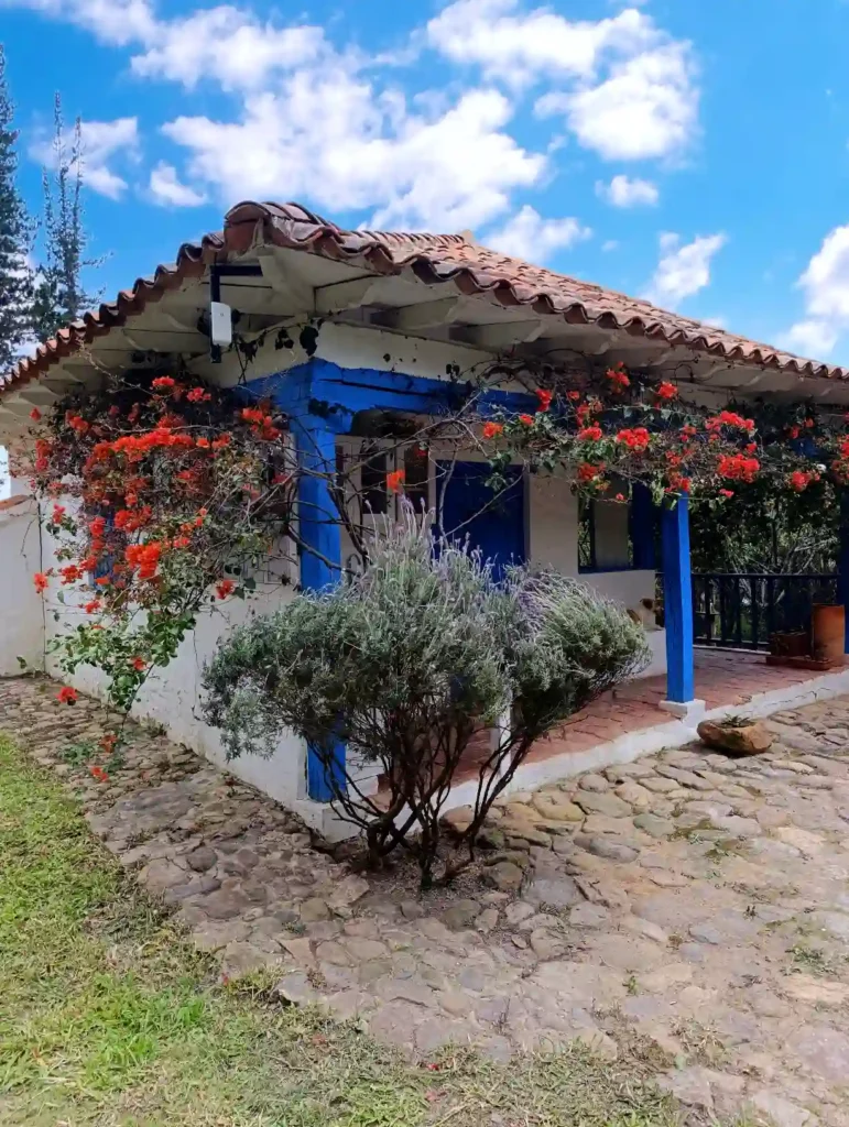 Casa de campo premium en Villa de Leyva: paredes blancas, tejado de teja y buganvillas rojas sobre un camino de piedra.