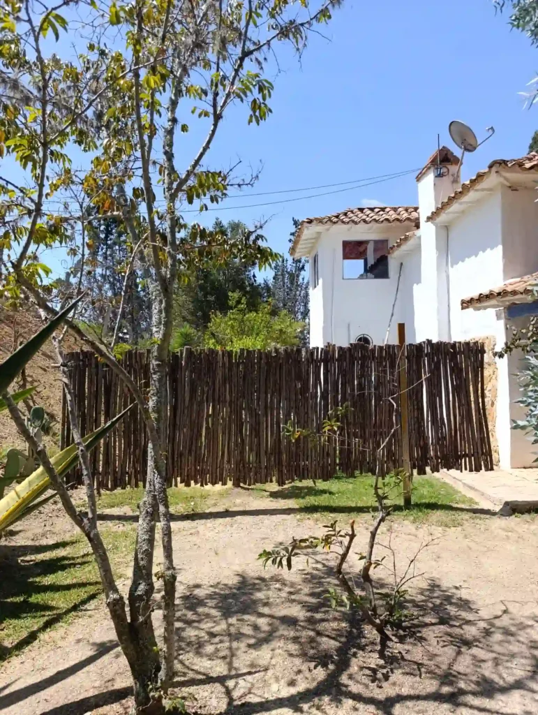 Vista rural de cabaña oteo en Villa de Leyva con techo de teja, valla de madera natural y vegetación. Ambiente seguro y privado.