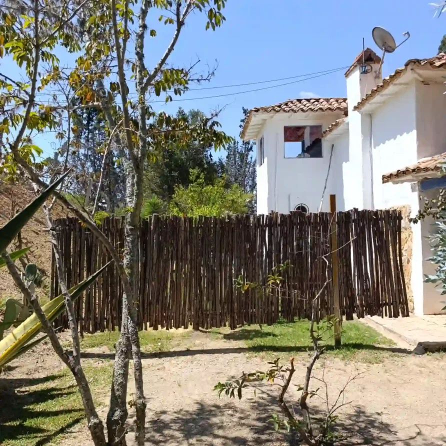 Vista rural de cabaña oteo en Villa de Leyva con techo de teja, valla de madera natural y vegetación. Ambiente seguro y privado.