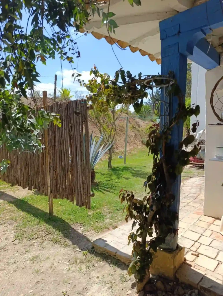 Cabaña rústica en Villa de Leyva con cerca de madera, jardín verde y pilar azul, evocando hospedaje campestre.