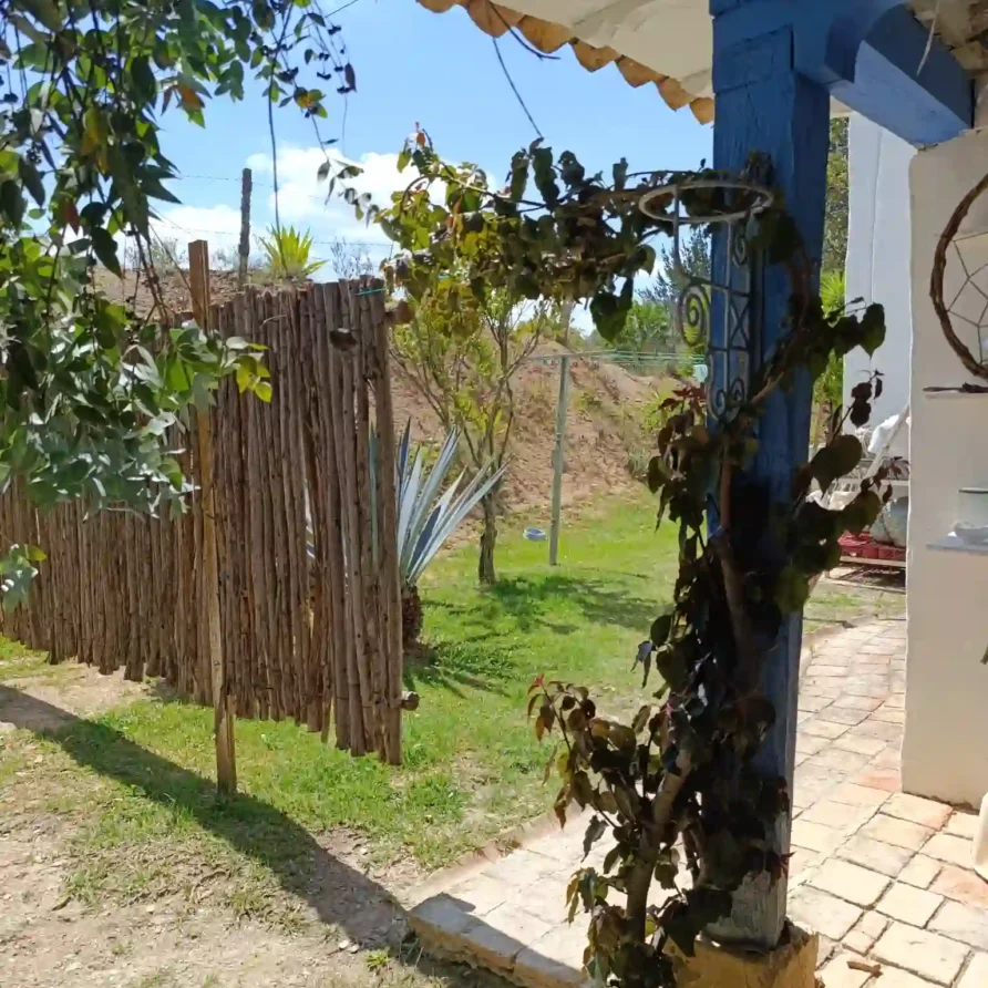 Cabaña rústica en Villa de Leyva con cerca de madera, jardín verde y pilar azul, evocando hospedaje campestre.